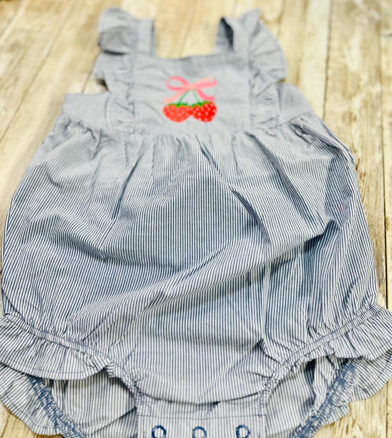 May include: Blue and white striped baby romper with a cherry applique. The romper features ruffled straps and leg openings. The cherry design has red cherries and a pink bow. The fabric has a seersucker texture.