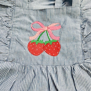 May include: Blue and white striped apron with a ruffled edge and straps. The apron features an embroidered design of two red strawberries with green stems and a pink bow.