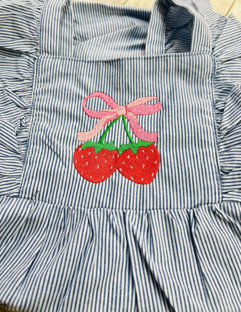 May include: Blue and white striped apron with a ruffled edge and straps. The apron features an embroidered design of two red strawberries with green stems and a pink bow.