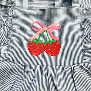 May include: Blue and white striped apron with a ruffled edge and straps. The apron features an embroidered design of two red strawberries with green stems and a pink bow.