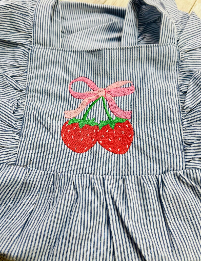 May include: Blue and white striped apron with a ruffled edge and straps. The apron features an embroidered design of two red strawberries with green stems and a pink bow.