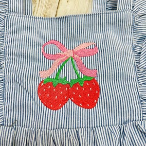 May include: Close-up of a blue and white striped apron with a red strawberry embroidery design. The strawberries have green stems and are connected by a pink bow. The apron has ruffled edges and straps.