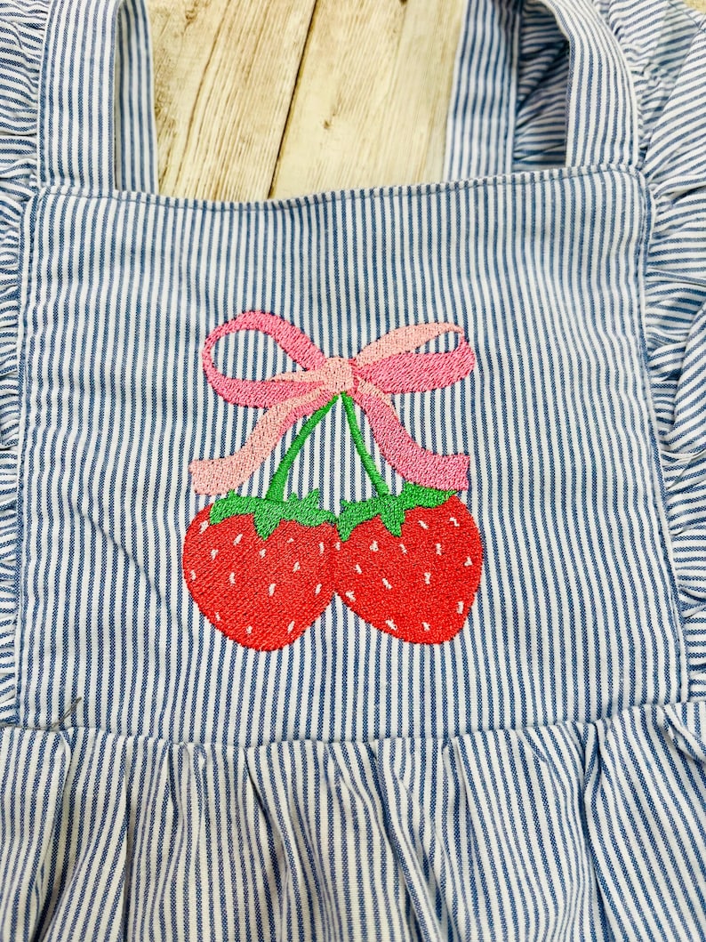 May include: Close-up of a blue and white striped apron with a red strawberry embroidery design. The strawberries have green stems and are connected by a pink bow. The apron has ruffled edges and straps.