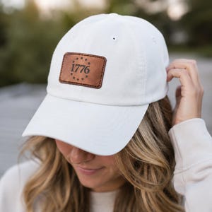 Può includere: Un cappellino da baseball bianco con una toppa rettangolare in pelle marrone sulla parte anteriore. La toppa presenta il numero 1776 in rilievo circondato da un cerchio di tredici piccole stelle. Il cappellino ha una visiera curva e una calotta.