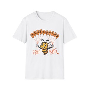 May include: A white short-sleeve t-shirt featuring a cartoon bee holding orange balloon-shaped letters that spell HOMECOMING. Below the bee, the text 2026 and KHS are printed in orange, accompanied by a small orange pencil graphic.