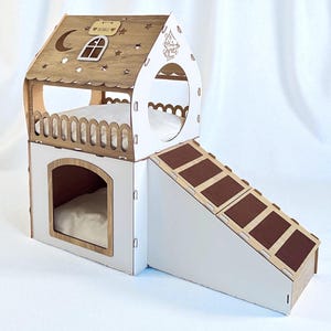 Peut inclure: Une maison pour chat à deux étages, blanche et boisée, avec une rampe. Le toit est orné d'étoiles, d'une lune et d'une fenêtre. Chaque niveau dispose d'un lit. La rampe a des marches marron.