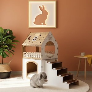 May include: A two-story wooden pet house with a white and natural wood finish, featuring a small staircase and a fenced balcony. A gray rabbit sits near the house on a cream-colored rug. A framed bunny silhouette hangs on the wall.