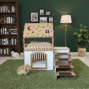 Op de afbeelding: Een houten dierenhuis met twee verdiepingen en een huisvormig ontwerp. De bovenste verdieping heeft een bed met een decoratieve balustrade en het dak heeft maan- en steruitsparingen. De onderste verdieping heeft een deur en een trap.