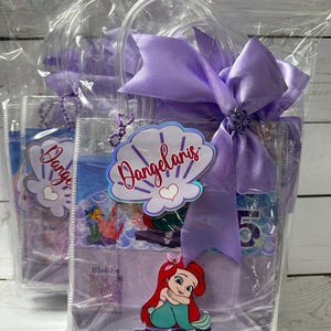 May include: Clear plastic gift bags with purple handles and bows. Each bag features a seashell-shaped tag with the name "Dangelaris" and a cartoon image of Ariel from The Little Mermaid. The bags are filled with party favors.