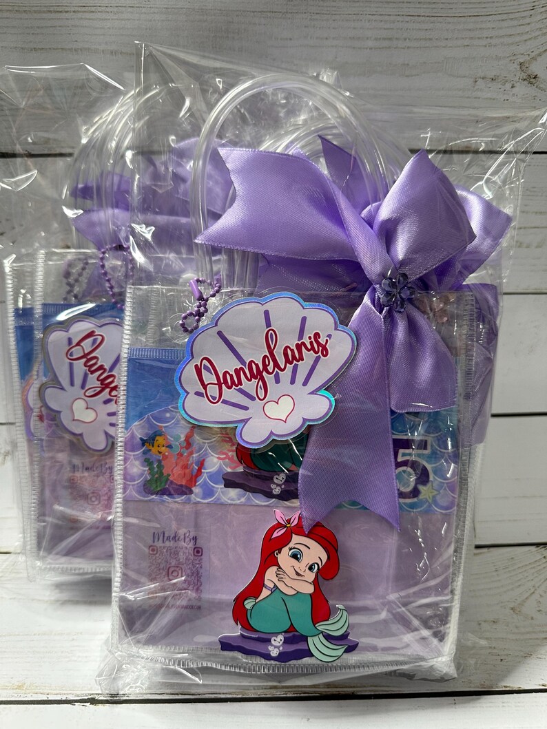 May include: Clear plastic gift bags with purple handles and bows. Each bag features a seashell-shaped tag with the name "Dangelaris" and a cartoon image of Ariel from The Little Mermaid. The bags are filled with party favors.