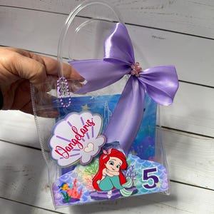 May include: A clear plastic gift bag with a purple satin bow and a decorative Ariel the Little Mermaid design. The bag includes the name "Dangelaris" and the number "5".