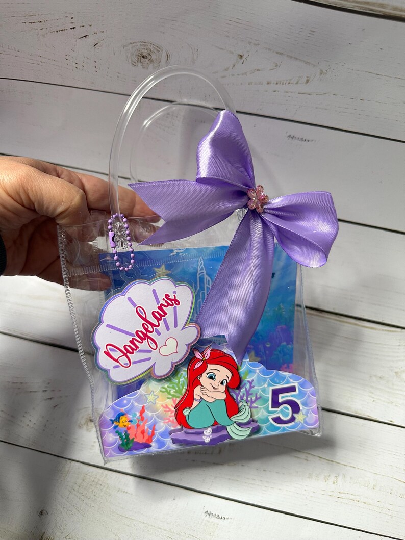 May include: A clear plastic gift bag with a purple satin bow and a decorative Ariel the Little Mermaid design. The bag includes the name "Dangelaris" and the number "5".