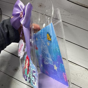 May include: Clear plastic tote bag with a purple bow and a charm. Inside is a card featuring Ariel from The Little Mermaid and the number 5. The card has an underwater scene with a castle and fish.
