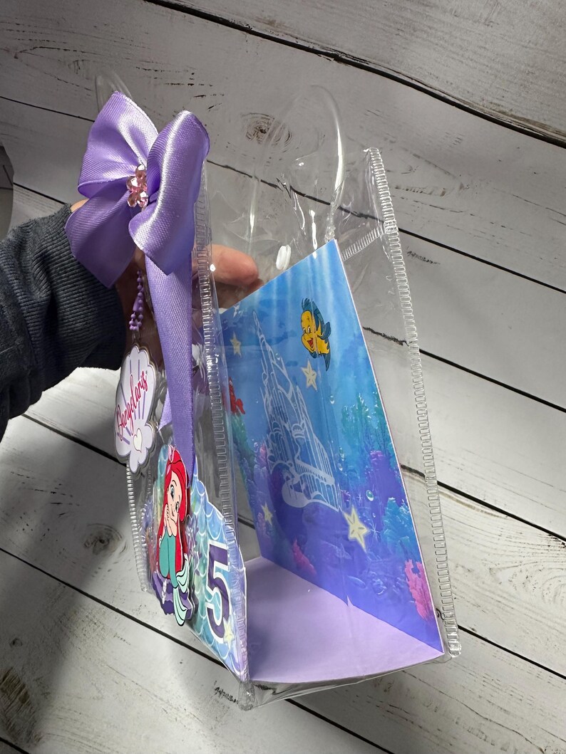 May include: Clear plastic tote bag with a purple bow and a charm. Inside is a card featuring Ariel from The Little Mermaid and the number 5. The card has an underwater scene with a castle and fish.