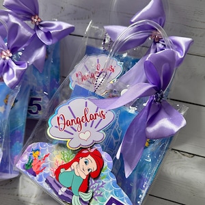 May include: Clear plastic favor bags with purple satin bows and handles. Each bag features a mermaid-themed design with the number 5. The bags have a personalized label with the name "Dangelaris".