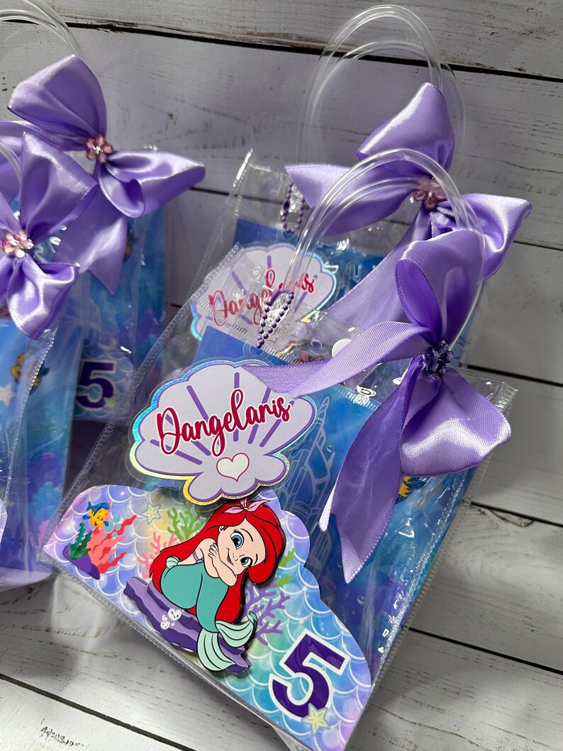 May include: Clear plastic favor bags with purple satin bows and handles. Each bag features a mermaid-themed design with the number 5. The bags have a personalized label with the name "Dangelaris".