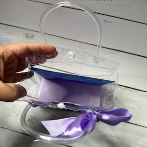May include: A clear plastic gift bag with a rounded handle and a lavender satin ribbon bow. The bag is partially open, revealing a lavender interior and a colorful insert.