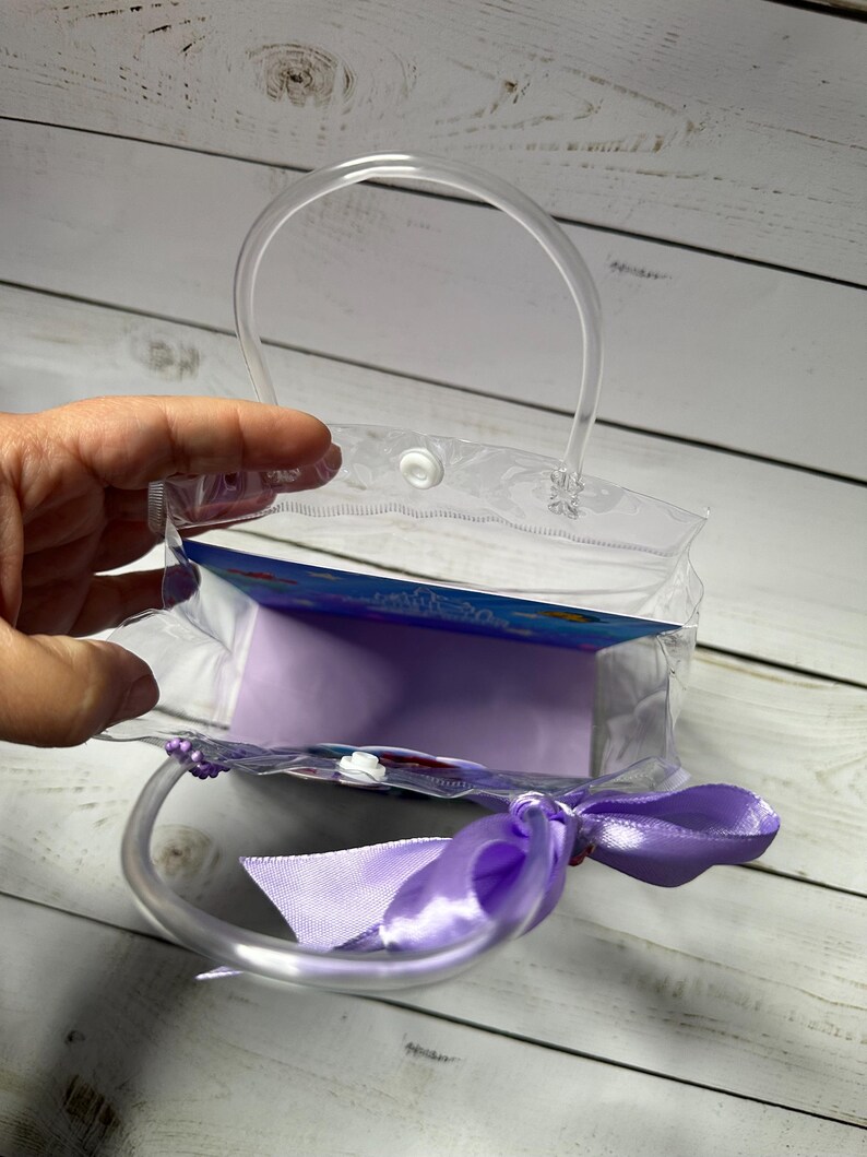 May include: A clear plastic gift bag with a rounded handle and a lavender satin ribbon bow. The bag is partially open, revealing a lavender interior and a colorful insert.