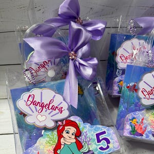 May include: Clear plastic favor bags with purple satin bows and the name "Dangelaris" on a seashell-shaped label. Each bag features a colorful image of Ariel from The Little Mermaid and the number 5.