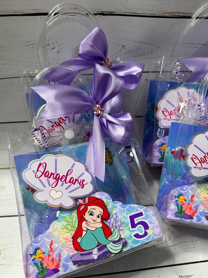 May include: Clear plastic favor bags with purple satin bows and the name "Dangelaris" on a seashell-shaped label. Each bag features a colorful image of Ariel from The Little Mermaid and the number 5.