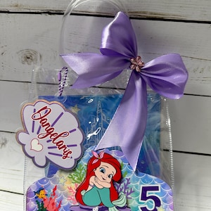 May include: A clear plastic gift bag with a purple bow and a decorative tag. The tag features the name "Dangelaris" in script, a seashell design, and a heart. The bag also includes a mermaid-themed cutout with the number "5".