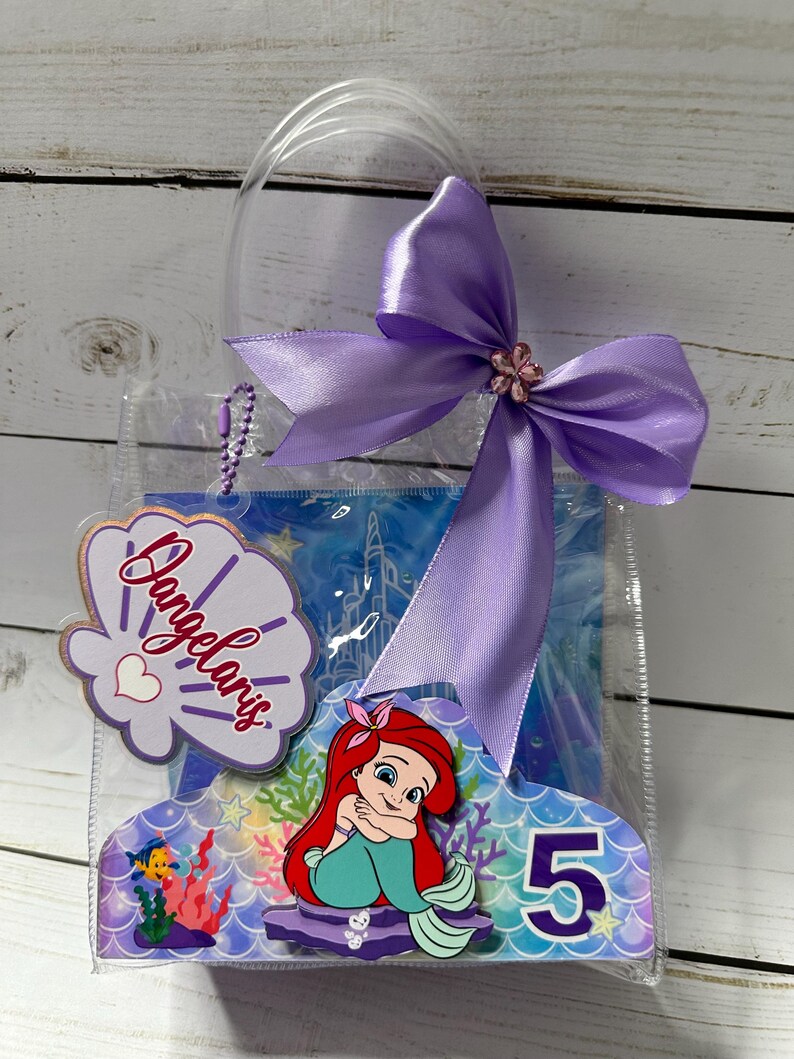 May include: A clear plastic gift bag with a purple bow and a decorative tag. The tag features the name "Dangelaris" in script, a seashell design, and a heart. The bag also includes a mermaid-themed cutout with the number "5".