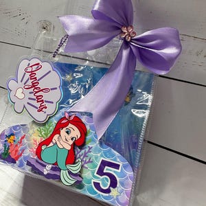 May include: Clear plastic gift bag with a purple satin bow and a decorative flower. The bag features a cartoon image of Ariel from The Little Mermaid, the number 5, and the name "Angelaris" on a seashell-shaped tag.
