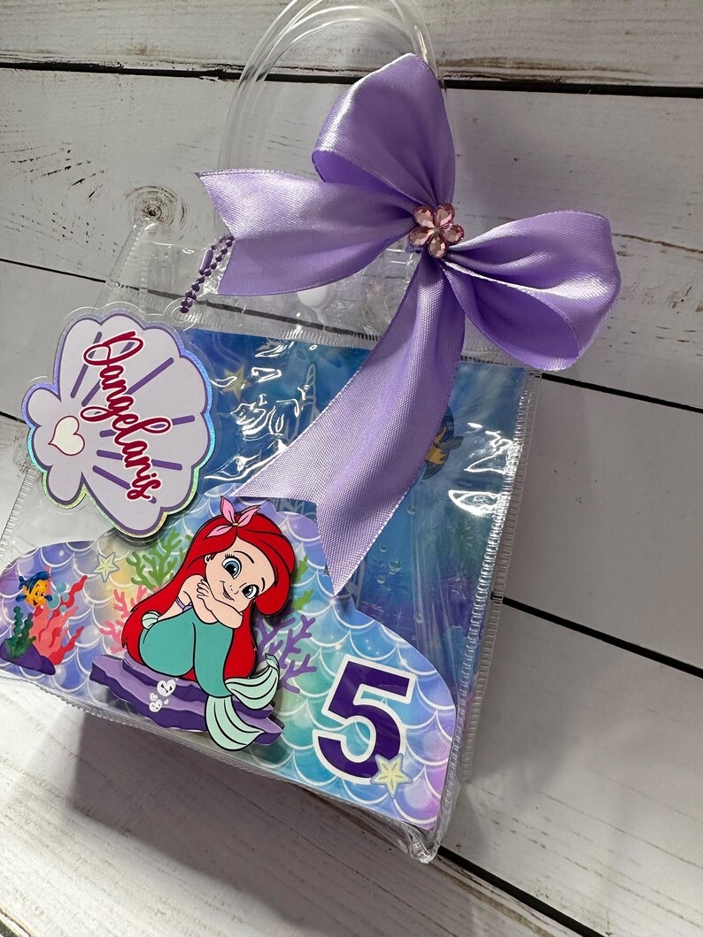 May include: Clear plastic gift bag with a purple satin bow and a decorative flower. The bag features a cartoon image of Ariel from The Little Mermaid, the number 5, and the name "Angelaris" on a seashell-shaped tag.