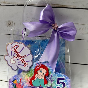 May include: Clear plastic gift bag with a purple bow and a decorative seashell-shaped tag that reads "Dangelaris". The bag features a cartoon image of Ariel the Little Mermaid, the number 5, and a blue background.