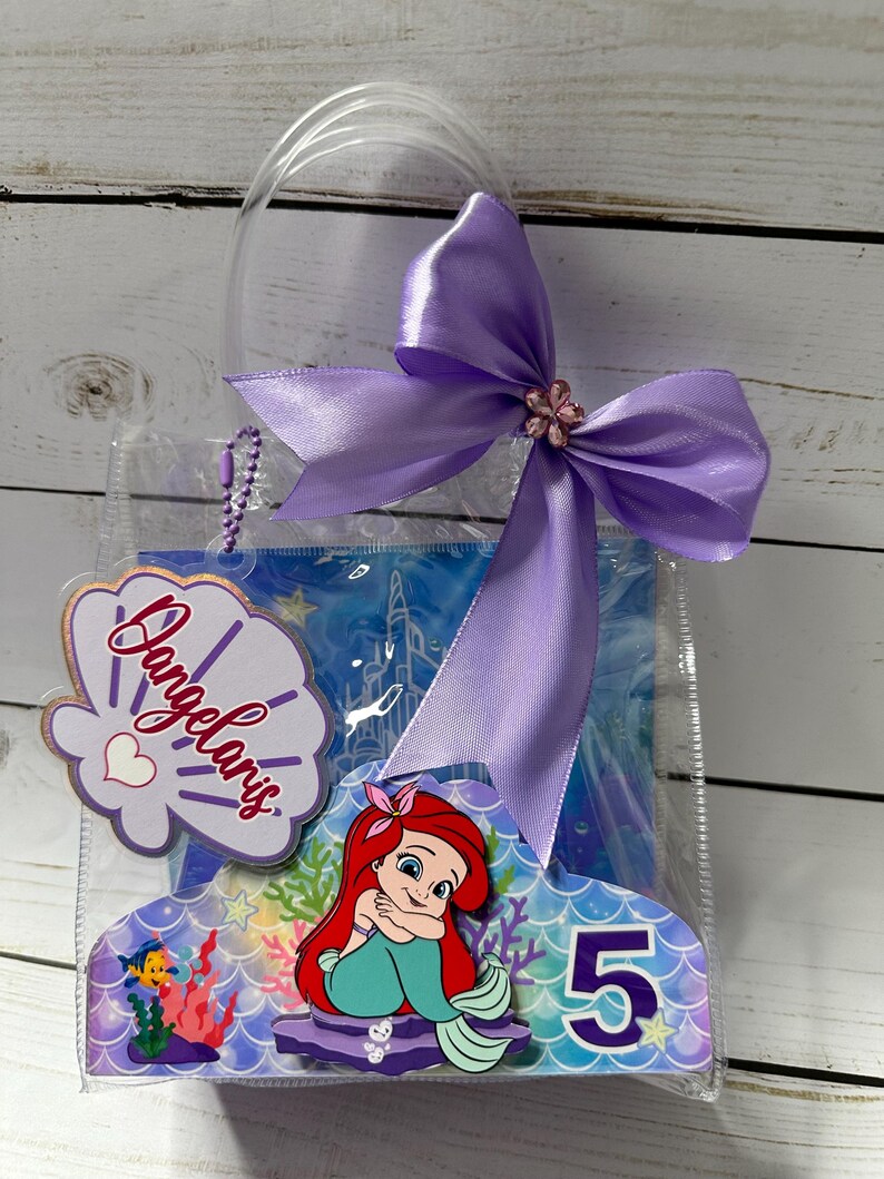 May include: Clear plastic gift bag with a purple bow and a decorative seashell-shaped tag that reads "Dangelaris". The bag features a cartoon image of Ariel the Little Mermaid, the number 5, and a blue background.