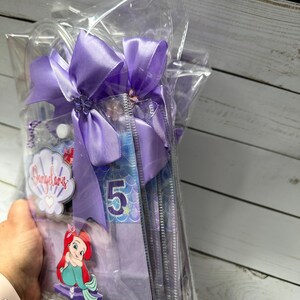 May include: A clear cellophane bag tied with a large lavender bow. Inside are several items, including a mermaid-themed decoration with the number 5, a seashell-shaped tag with text, and a cartoon image of Ariel.