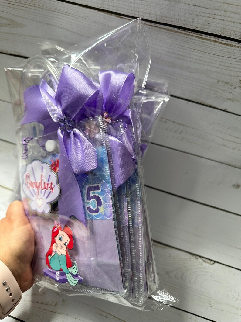 May include: A clear cellophane bag tied with a large lavender bow. Inside are several items, including a mermaid-themed decoration with the number 5, a seashell-shaped tag with text, and a cartoon image of Ariel.