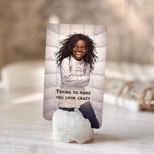 Pu&ograve; includere: Una carta rettangolare con un'illustrazione di una persona in una camicia di forza bianca e il testo "TRYING TO MAKE YOU LOOK CRAZY". La carta &egrave; sostenuta da un cristallo bianco.