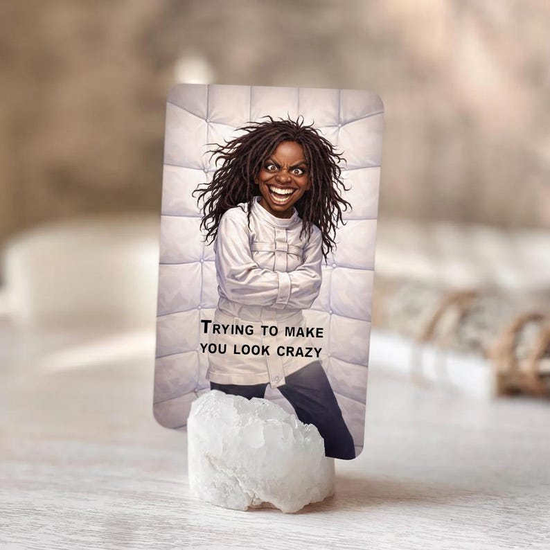 Pu&ograve; includere: Una carta rettangolare con un'illustrazione di una persona in una camicia di forza bianca e il testo "TRYING TO MAKE YOU LOOK CRAZY". La carta &egrave; sostenuta da un cristallo bianco.