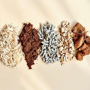 May include: Five distinct piles of organic materials arranged in a row, including light beige wood shavings, dark brown soil-like particles, small grey cylindrical pellets, light tan wood chips, and coarse brown fibrous coconut husk chunks.