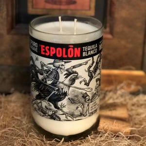 May include: A white candle in a clear glass with two wicks. The candle's label features the text "Espolòn Tequila Blanco" and a black and white illustration of skeletons. The candle is surrounded by a neutral background.