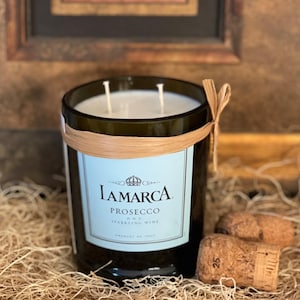 May include: A glass candle jar with a brown glass bottle base and a light blue label with the text "LAMARCA PROSECCO D.O.C. SPARKLING WINE" and a gold crown. The jar is wrapped with a brown bamboo band and has two white wicks. The candle is sitting on a bed of brown straw.