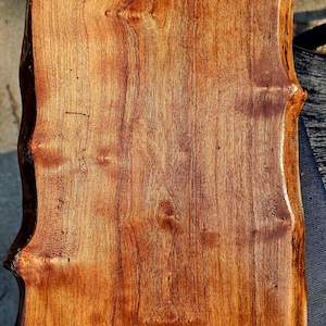 Puede incluir: Una tabla de cortar rectangular de madera con borde natural. La tabla tiene un color marrón rojizo intenso con vetas y nudos de madera visibles. Los bordes son ligeramente irregulares, lo que le da un aspecto rústico. La superficie es lisa y parece estar acabada.