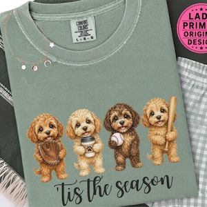 May include: A sage green t-shirt featuring a graphic of four brown puppies. One puppy holds a baseball glove, one holds a coffee cup, one holds a baseball, and one holds a wooden bat. The text 'Tis the season' is printed in black below the puppies.
