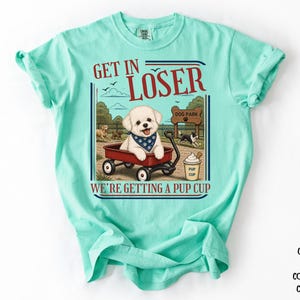 Peut inclure: T-shirt de couleur menthe crayeuse avec l'inscription "GET IN LOSER WE'RE GETTING A PUP CUP". Le graphisme représente un chien blanc dans un chariot rouge, un panneau de parc pour chiens et une "pup cup".