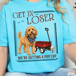 May include: A light blue t-shirt featuring a graphic of a golden-coloured dog wearing a blue bandana with white paw prints. Next to the dog is a red wagon and a cup with whipped cream. The text reads GET IN LOSER WE'RE GETTING A PUP CUP.
