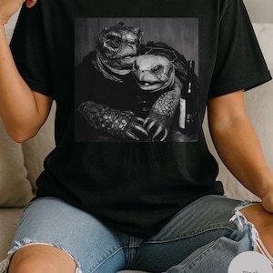 May include: Black t-shirt with a grayscale image of two turtles embracing, and a wine bottle. The image is centered on the shirt. The shirt is worn by a person wearing blue jeans.