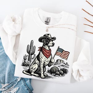 May include: White sweatshirt featuring a graphic of a dog in a cowboy hat and bandana, holding an American flag. The design includes cacti and a desert landscape. The sweatshirt is made of a soft material.