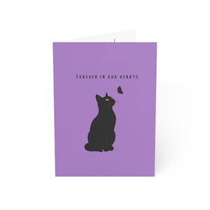 May include: A lavender greeting card featuring a black cat sitting and looking up at a small black butterfly. The text "FOREVER IN OUR HEARTS" is printed in black capital letters above the cat. The design is centred on the front of the card.