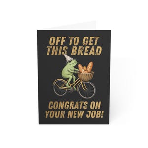 May include: A black greeting card featuring a green frog wearing a pointed hat, riding a yellow bicycle with a basket full of bread. The text reads OFF TO GET THIS BREAD and CONGRATS ON YOUR NEW JOB! in a distressed, light-colored font.