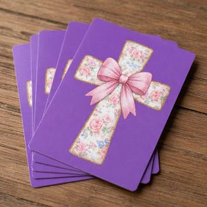 May include: A stack of purple cards featuring a floral cross design with a pink bow. The cross is decorated with roses and other flowers, and outlined in gold. The cards are stacked on a wooden surface.