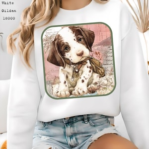 May include: White Gildan sweatshirt featuring a graphic of a puppy with brown and white spots, wearing a collar and holding a leaf. The graphic is set against a pink and floral background, framed by a green border.