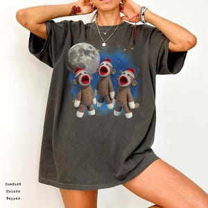 May include: Dark grey t-shirt featuring a graphic of three sock monkey dolls with open mouths, wearing red hats, and standing in front of a full moon and a blue starry background. The shirt has the words "Comfort Colors Pepper" printed on the bottom.