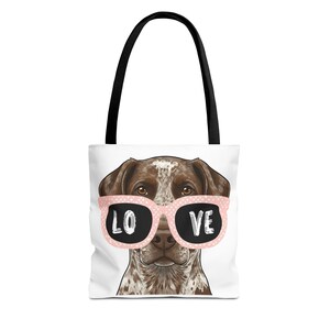 May include: White tote bag featuring a brown and white dog wearing pink sunglasses with the word "LOVE" in black. The bag has black handles and a white background.