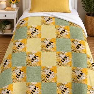 Peut inclure: Une courtepointe en patchwork composée de carrés alternés jaune, vert sauge et blanc. Les carrés à motifs présentent des illustrations de bourdons, de rayons de miel et de petites tournesols. Le couvre-lit recouvre un cadre de lit blanc.